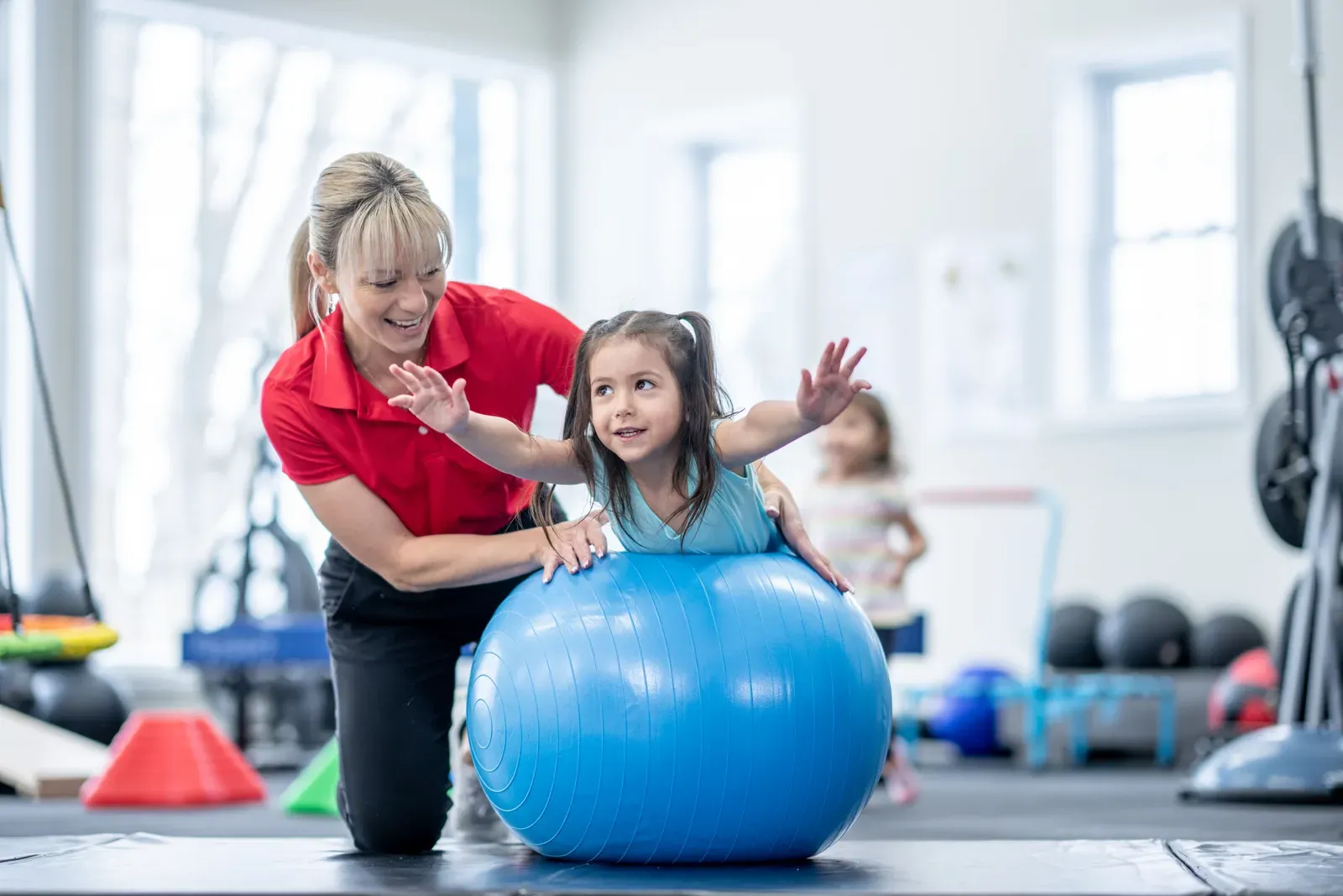 A therapist helping a young girl balance on an exercise ball in a bright gym