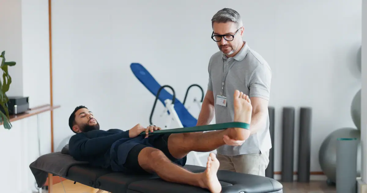 Physiotherapist helping a patient stretch a leg with a resistance band on a clinic table, a setting for blood flow restriction work