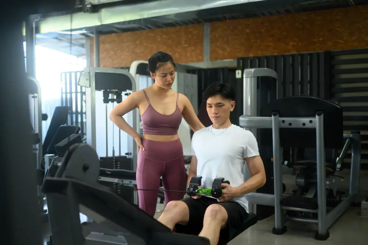 Female fitness coach supervising a male client on a seated cable row machine, equipment to weigh in a rented room