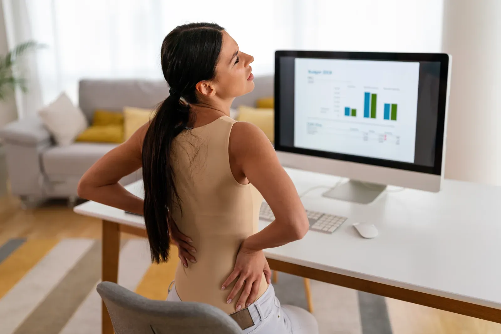 A woman at a desk computer holding her aching lower back