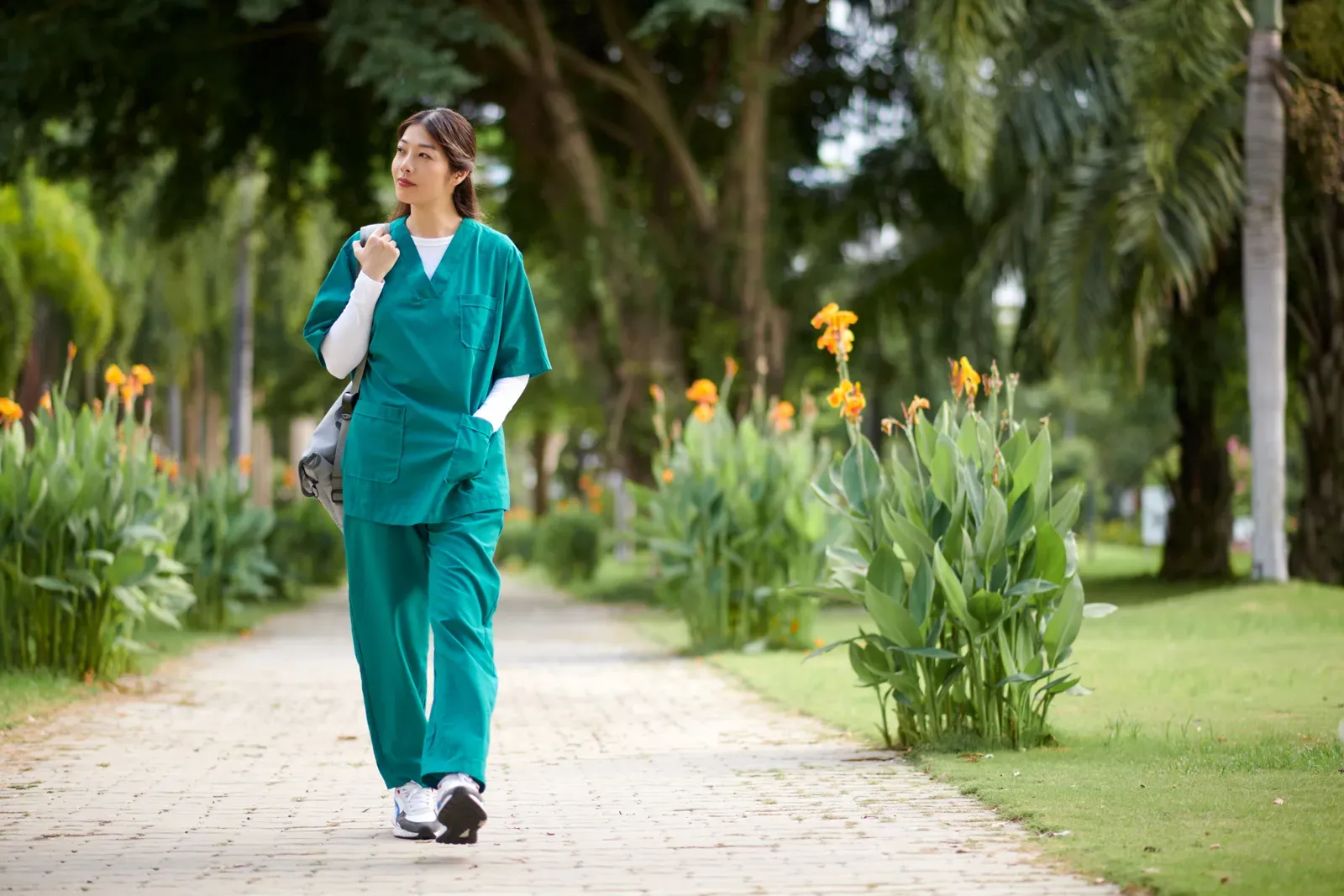 Nurse in scrubs with a shoulder bag walking along a tree lined path