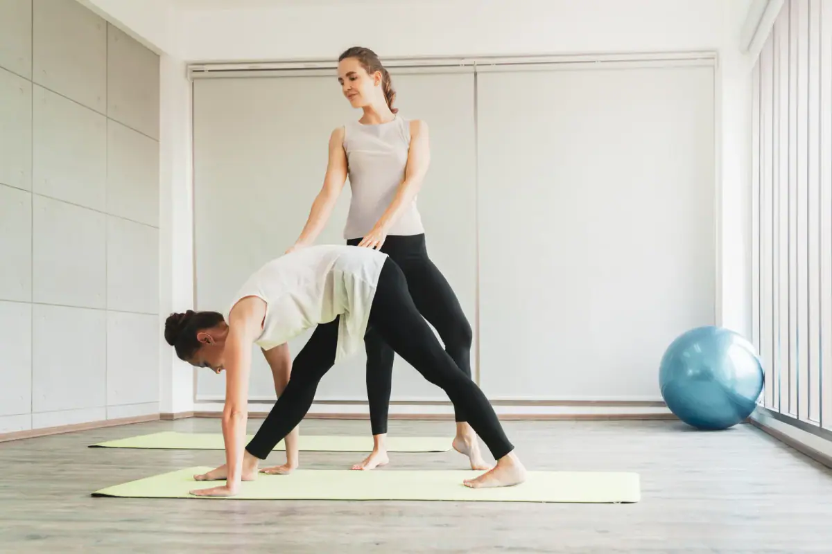 Trainer coaching a middle aged woman in a yoga class at a studio, a role a trainer might hire an assistant for