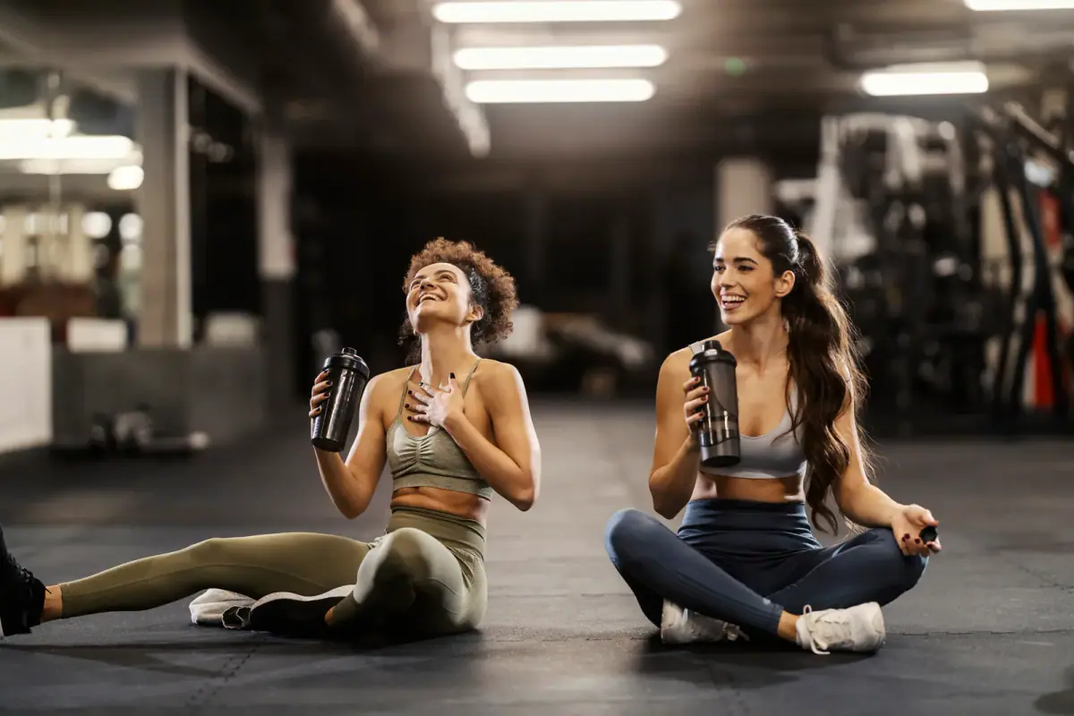 Two happy sporty friends sitting on a gym floor drinking water during a break, the kind of referral a client credit can reward