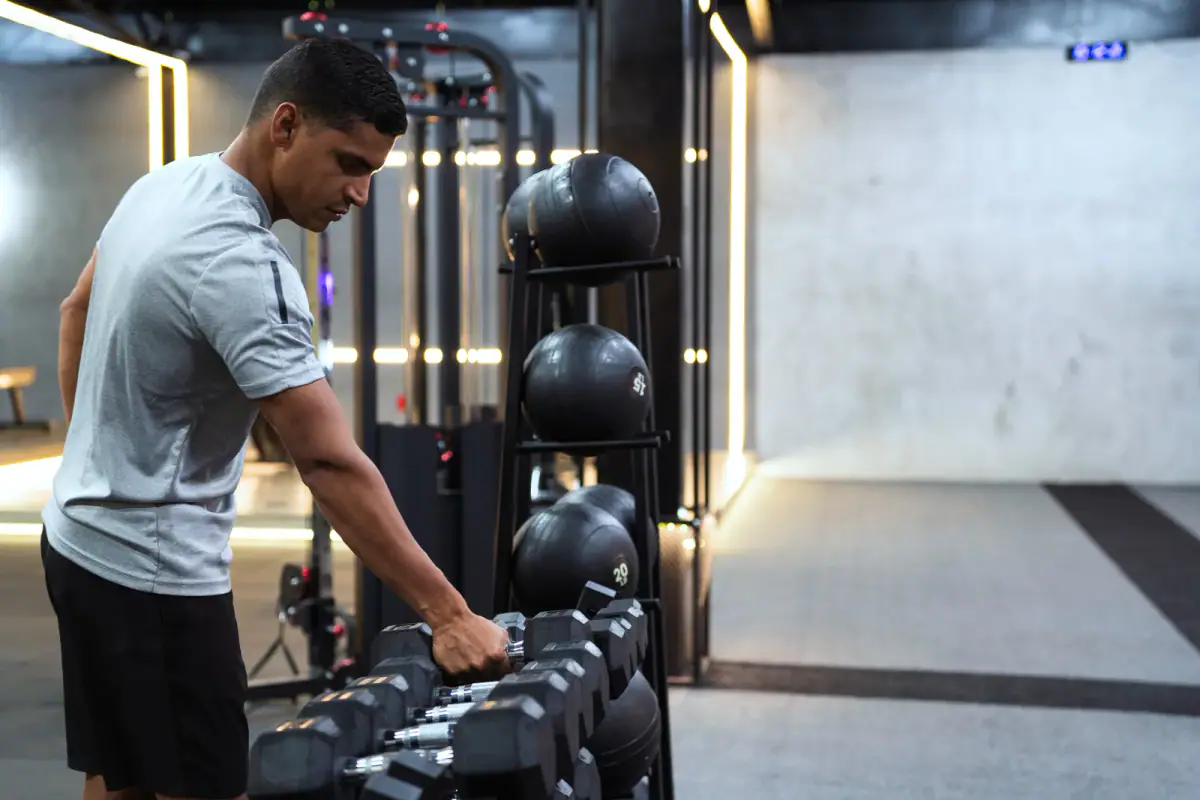 Young man selecting a dumbbell from the rack to prepare for a strength session, a gym routine to wind down when leaving a rented room