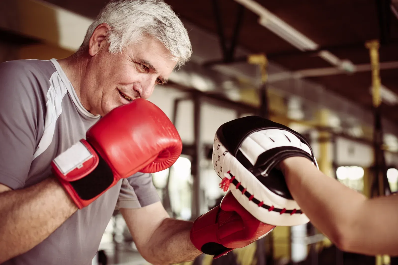 A senior man in boxing gloves working a punch mitt held by a trainer