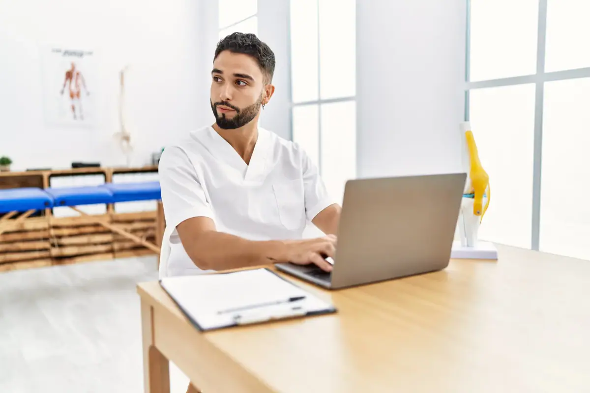 Young physiotherapist using a laptop at a clinic, a role to compare in physical therapist income, cash practice vs employed
