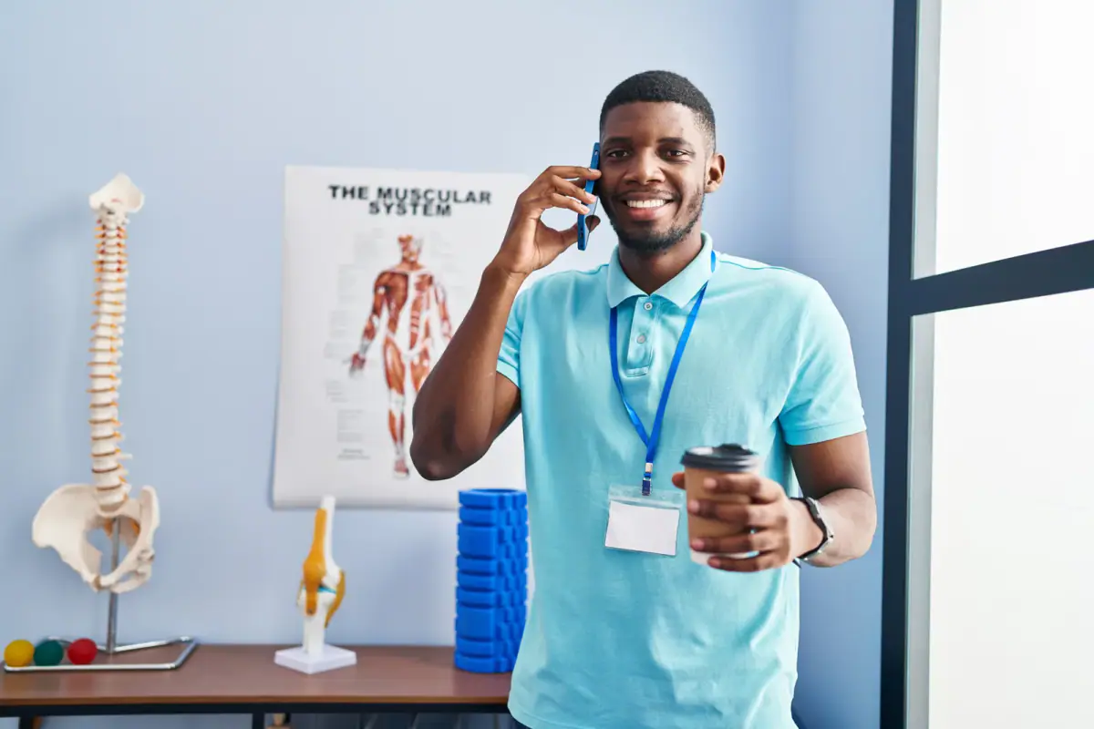 A physiotherapist talking on a smartphone with a coffee at a rehab clinic, like a call to update a client's doctor