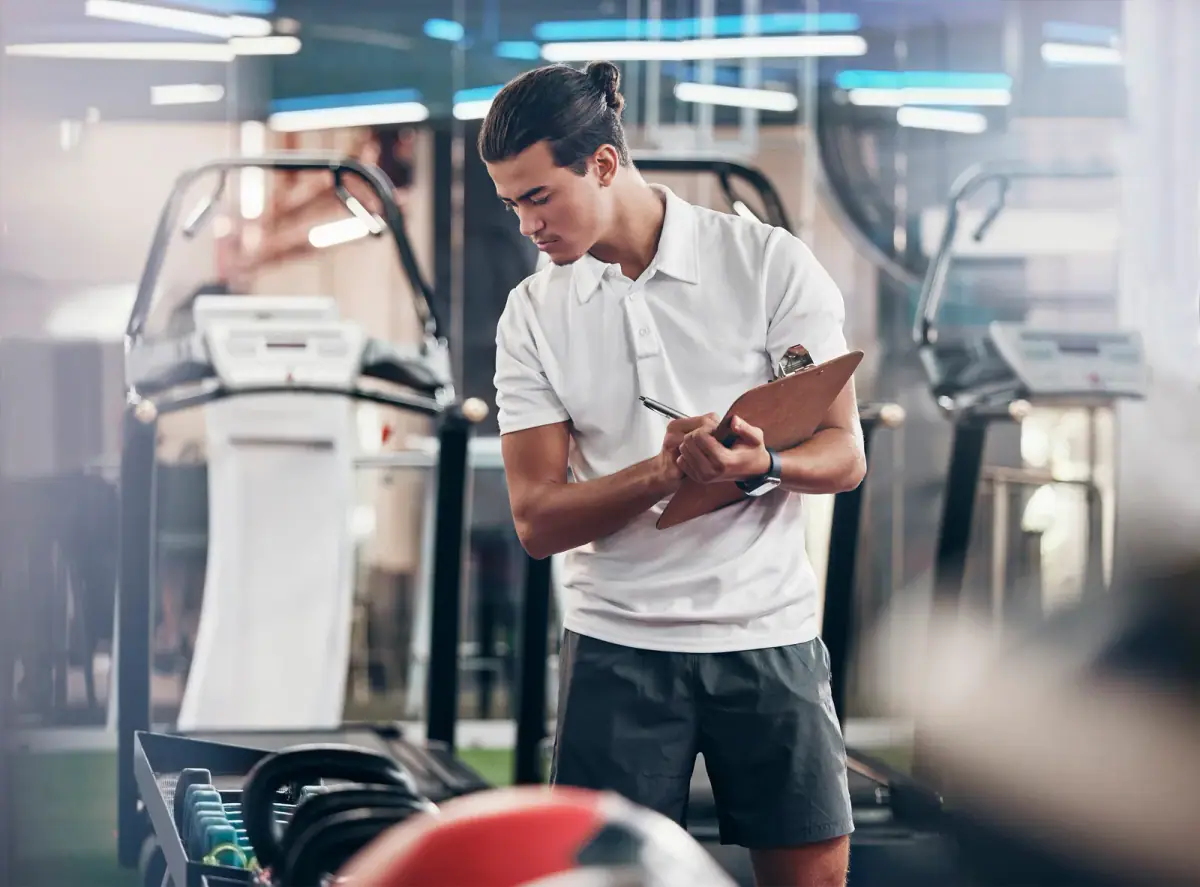 A manager with a clipboard checking equipment in a gym and writing notes, relevant to standard operating procedures