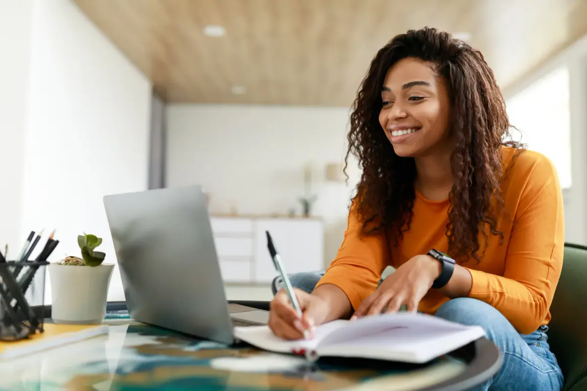 Black woman at a desk using a computer and writing in a notebook, a planning step for the startup costs of a solo trainer