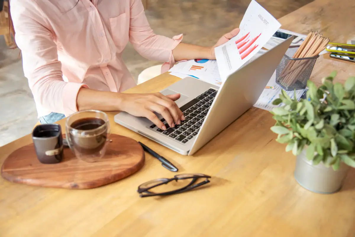 A woman working at a home office desk on a laptop with financial charts, relevant to a steady paycheck from irregular income