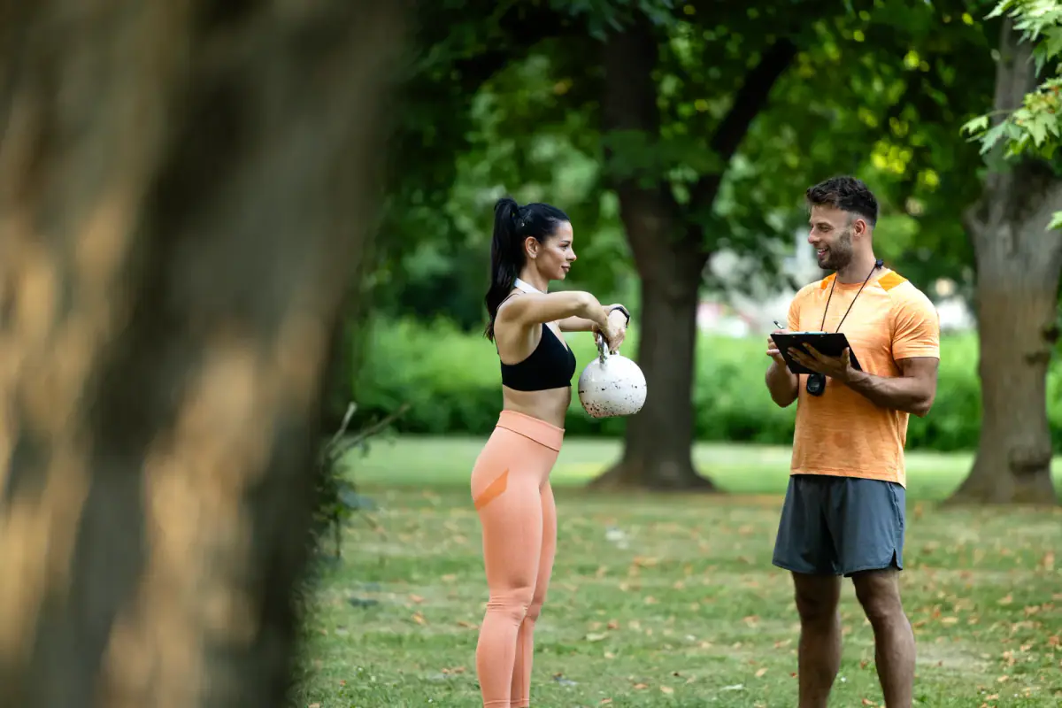 A fitness instructor evaluating a client's progress as a woman exercises with weights outdoors, relevant to training clients while employed