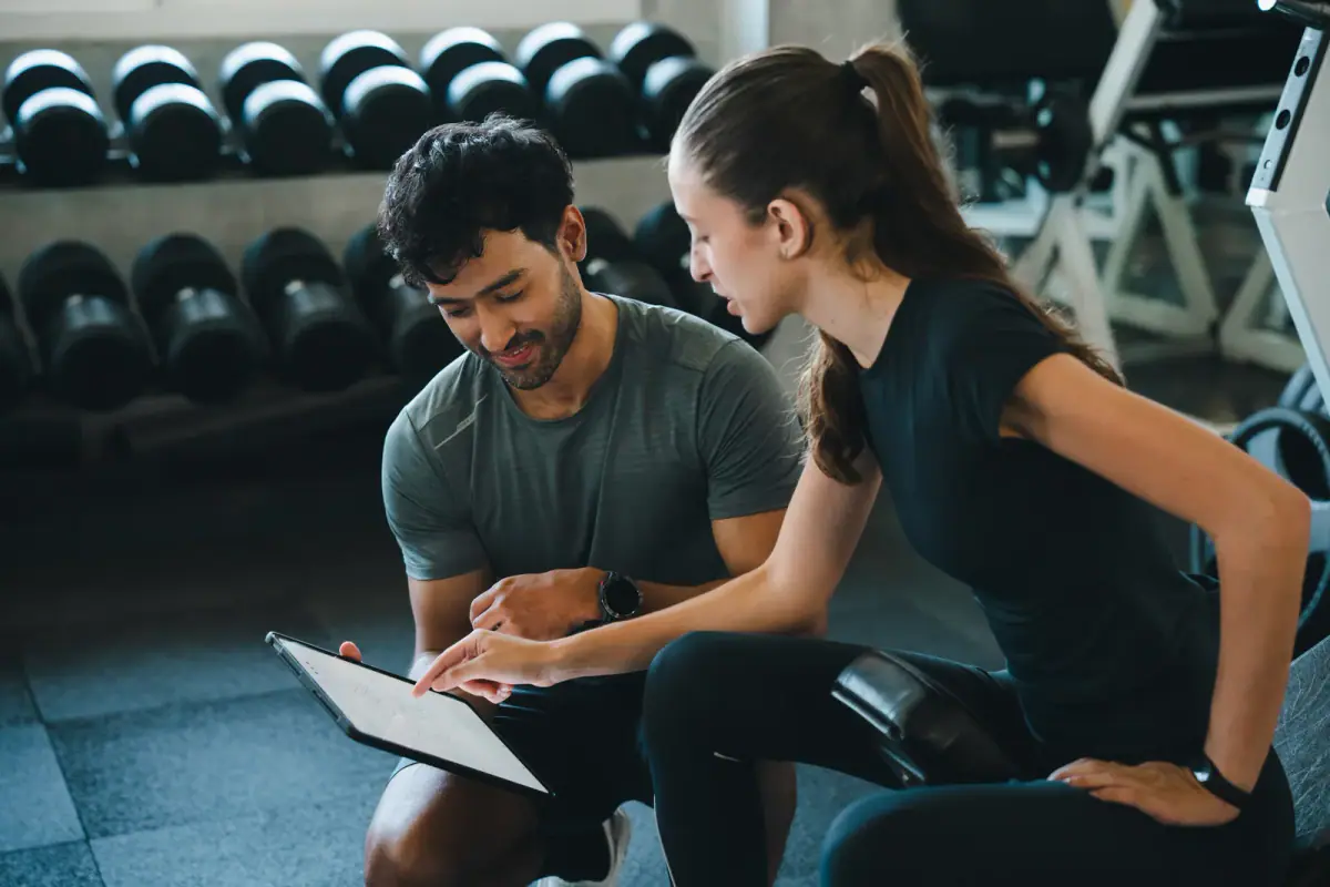 A personal trainer, a woman and a man in a workout consultation with a clipboard, relevant to two trainers splitting one suite