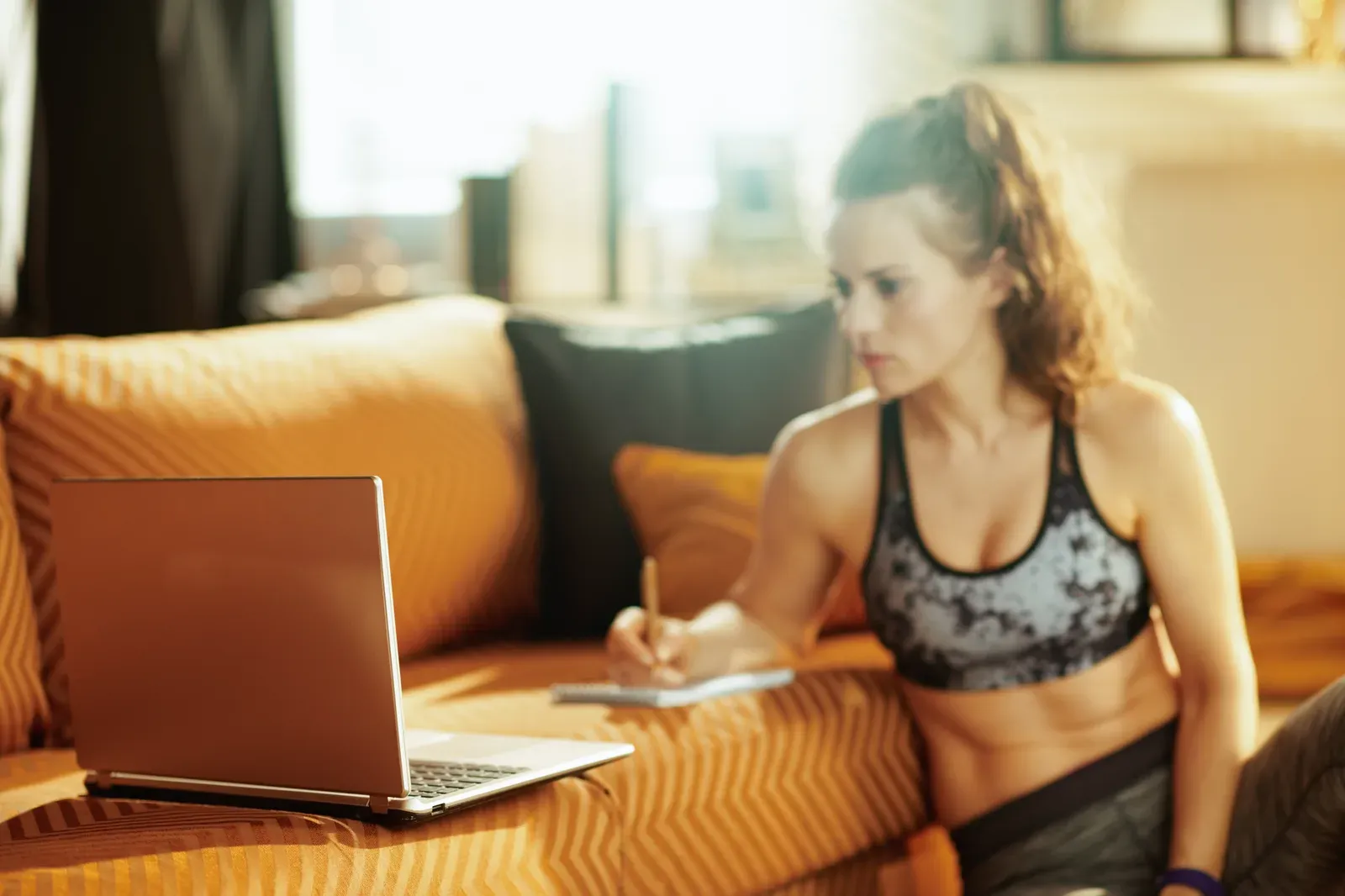 Woman planning workouts in a notebook beside an open laptop at home