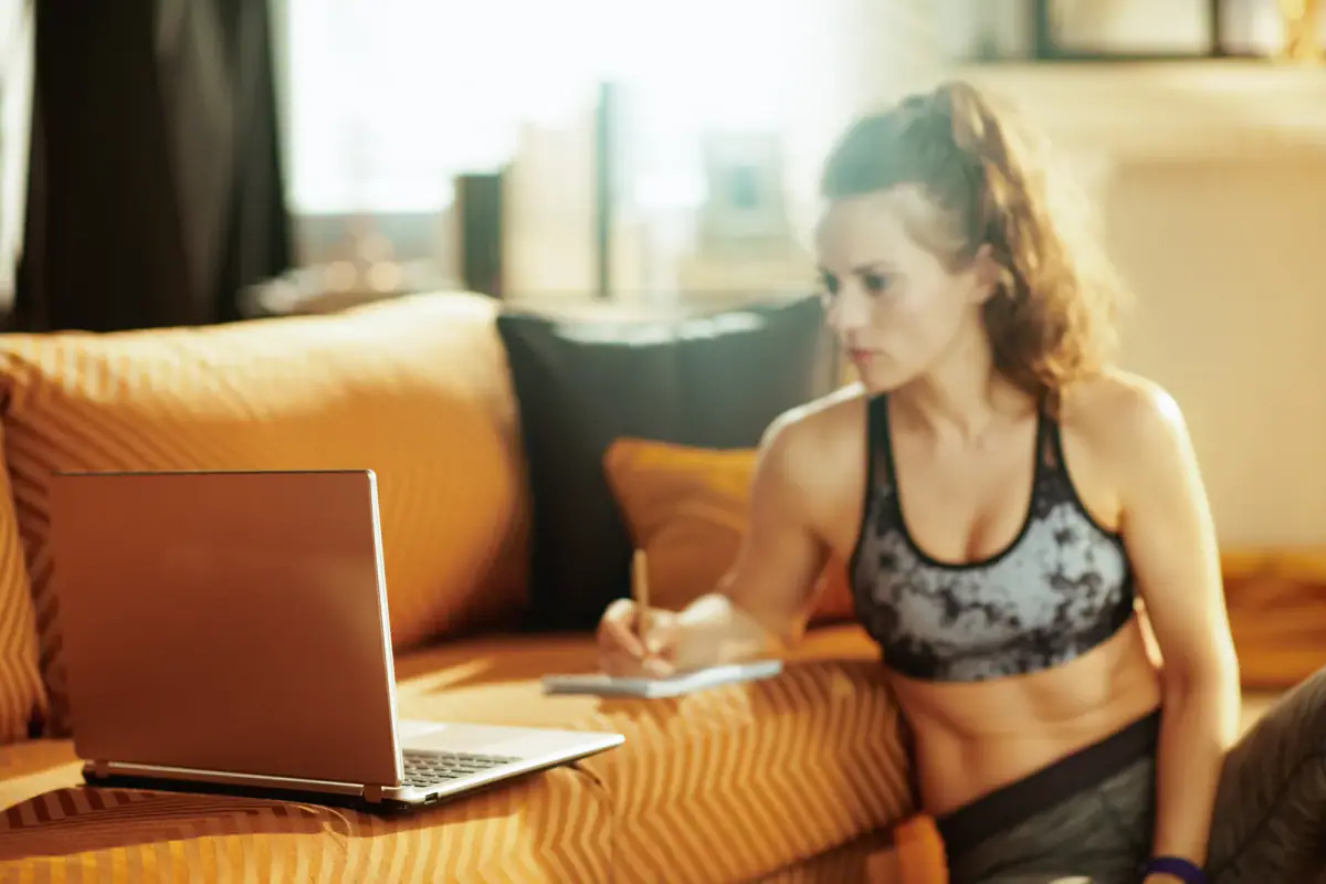 Woman planning workouts in a notebook beside an open laptop at home